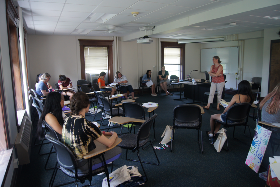 Encuentro participants seated in a classroom, facing a lecturer. There is a painting of different-colored handprints.