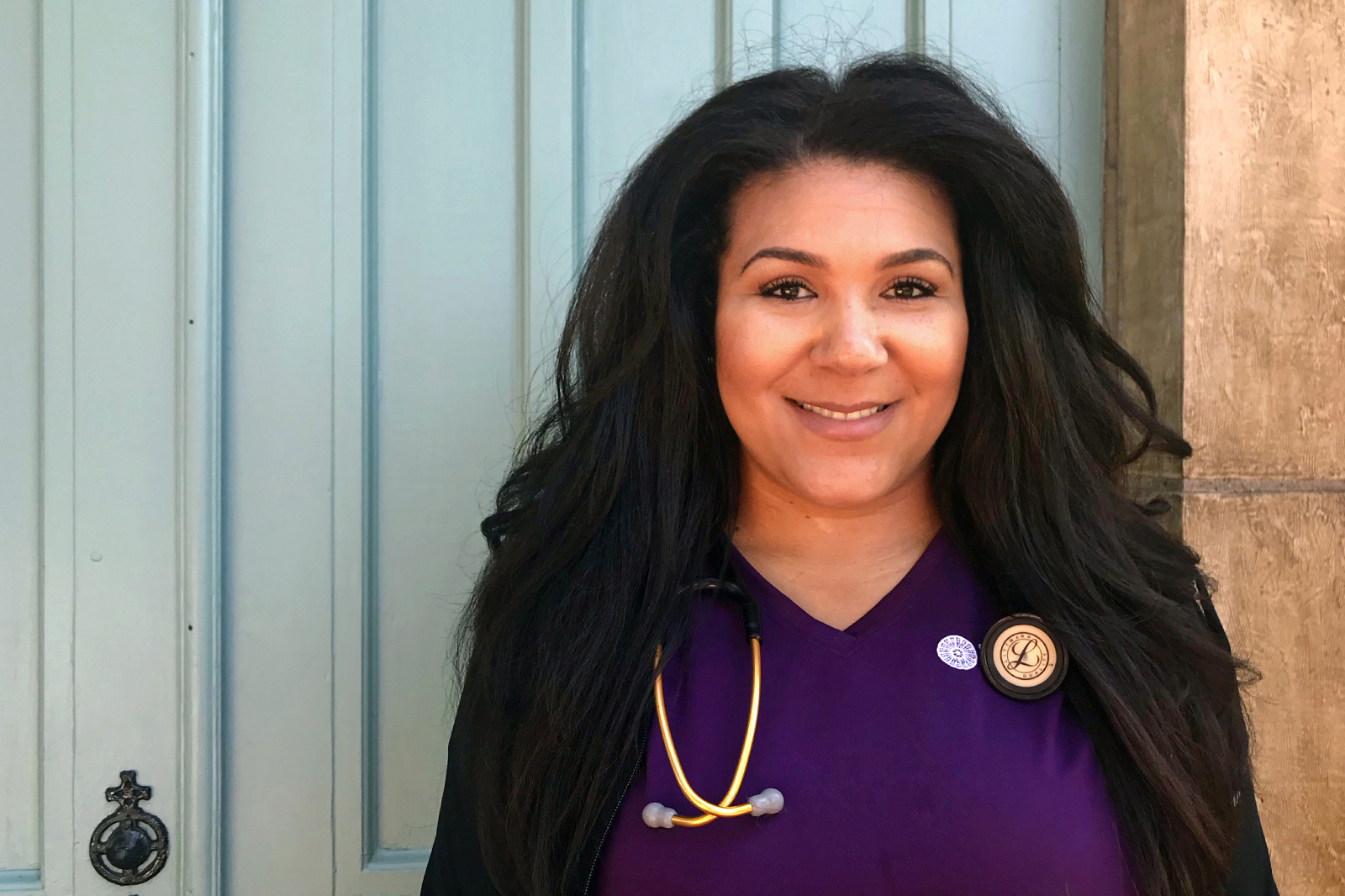 Photo of St. Catherine University undergraduate nursing program alumna Danyel Downs ’19, RN, CCRN, PHN, (standing in front of blue doors wearing purple scrubs and a stethoscope) won the Minnesota March of Dimes “Rising Star” award.