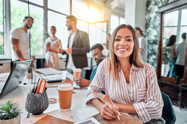 A smiling public relations specialist sits at an office desk working, with co-workers visible in the background.