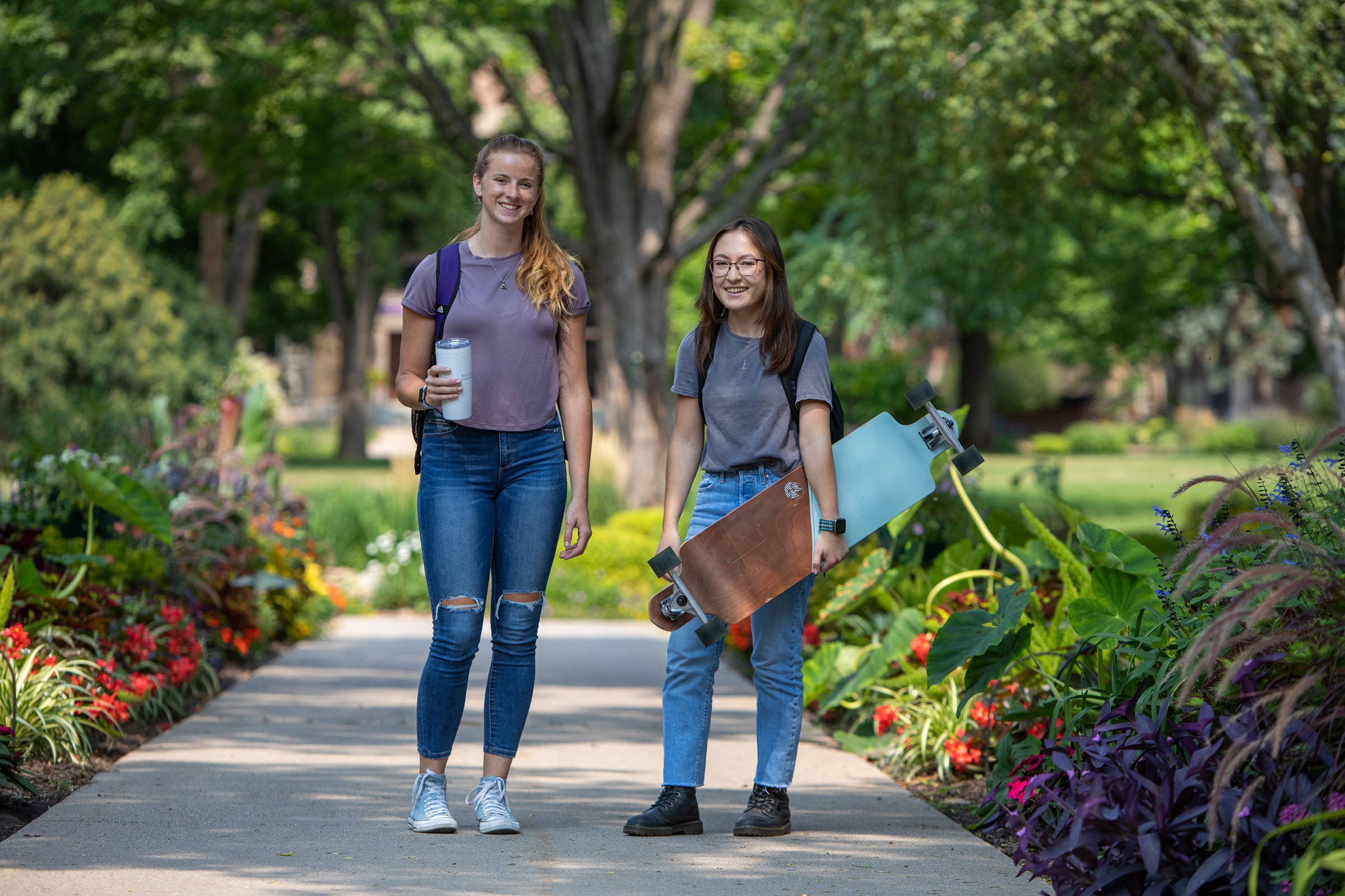 Two students, one with a coffee and another with her skateboard, standing alongside St. Kate's summer greenery