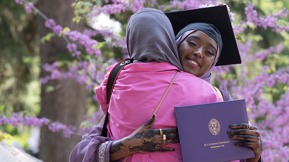 A new graduate in a cap and gown embraces someone in a hug. 