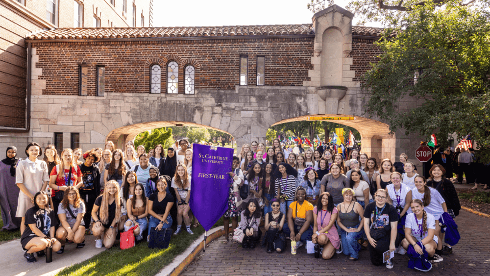 A large group of people gathers outside with a purple banner that reads "First Year"