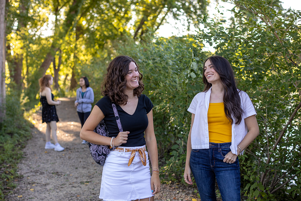 Four students walking on a wooded trail by a lake, talking and smiling.