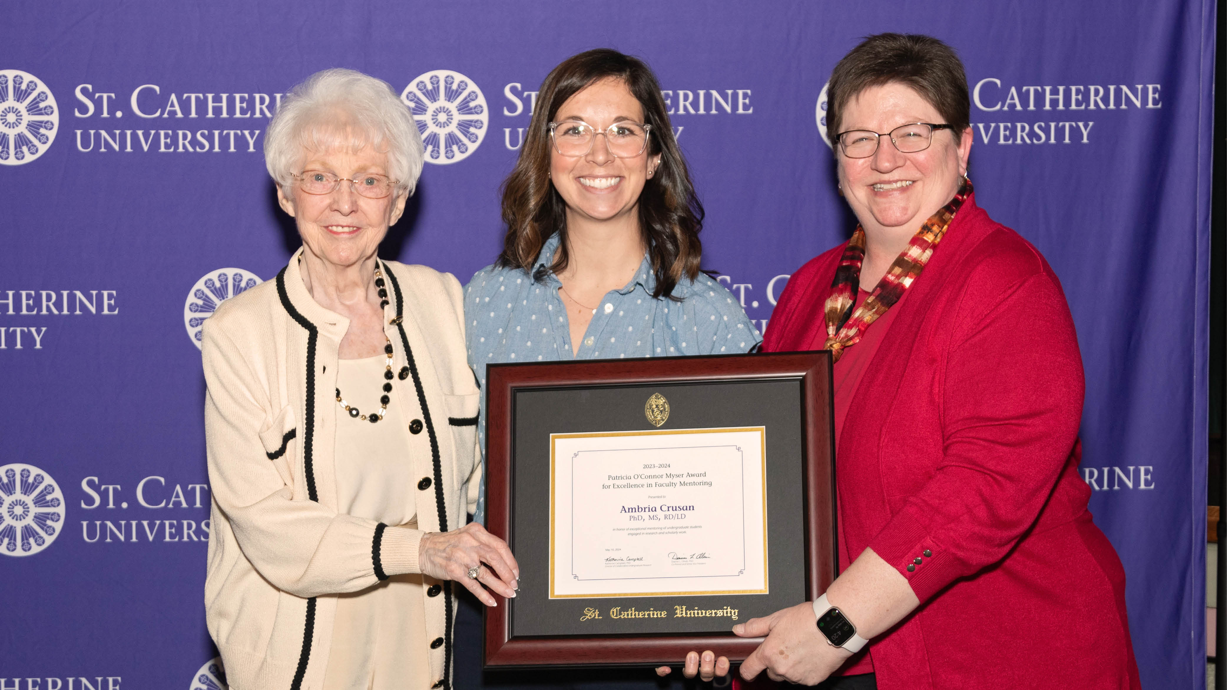 Three people stand in front of a purple background and hold a framed Patricia O’Connor Myser Award for Excellence in Faculty Mentoring