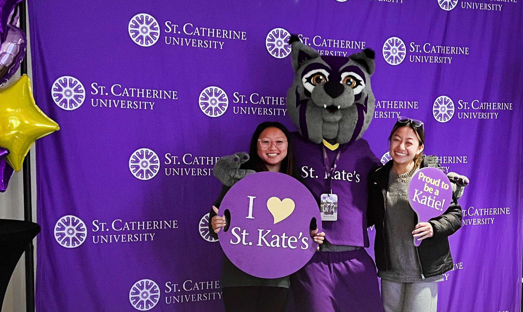 Two students, on either side of a Wildcat mascot, stand in front of a purple backdrop and hold signs that say "I love St. Kate's" and "Proud to be a Katie!"