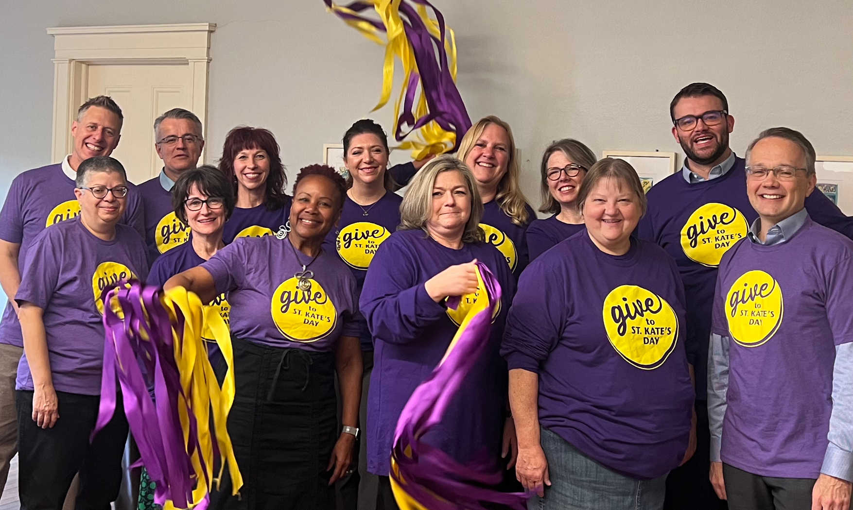 A group of a dozen people wearing purple "Give to St. Kate's Day" shirts smile and wave ribbons.