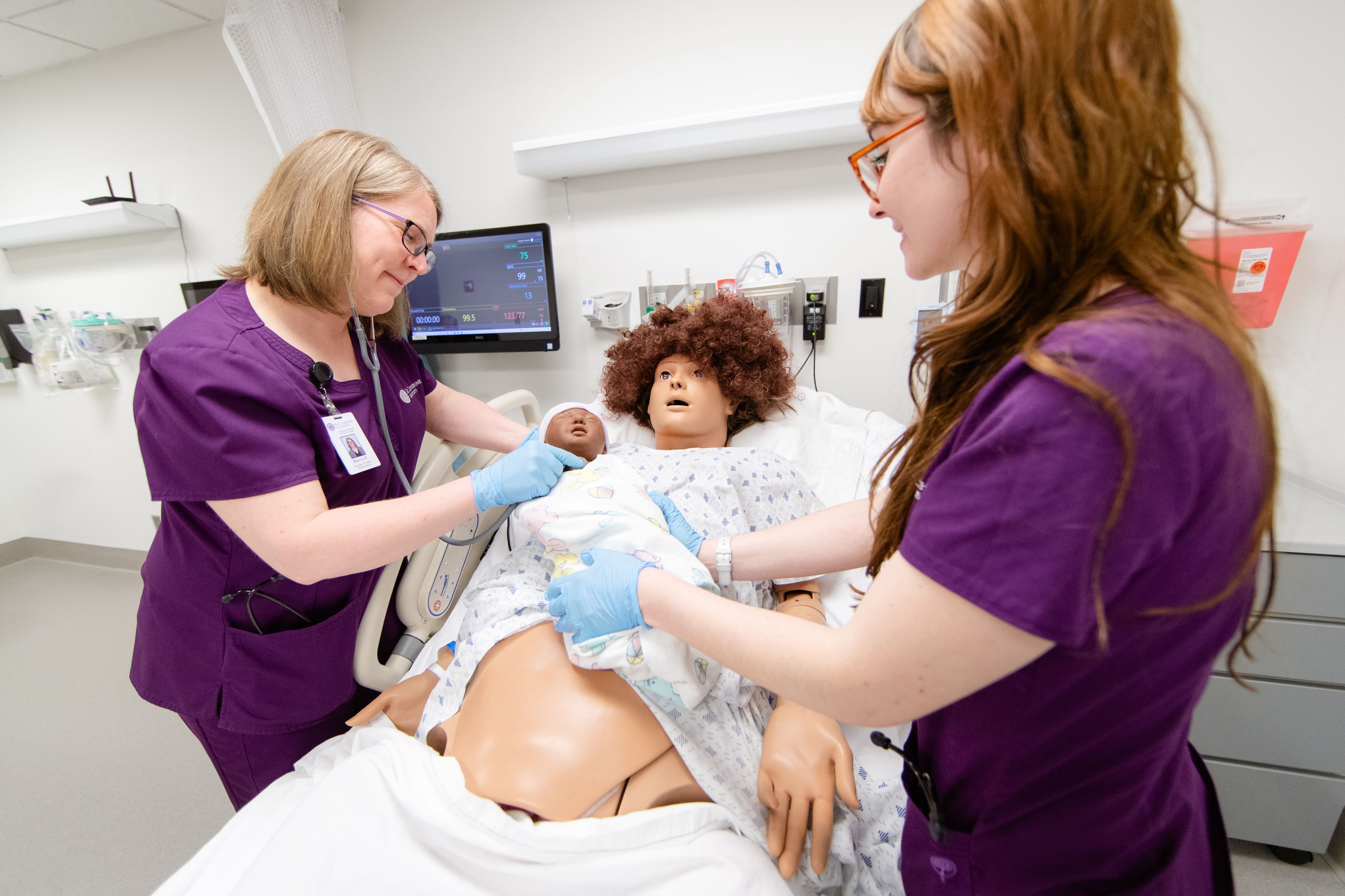 Two nursing student in purple scrubs working on a mannequin in a classroom 