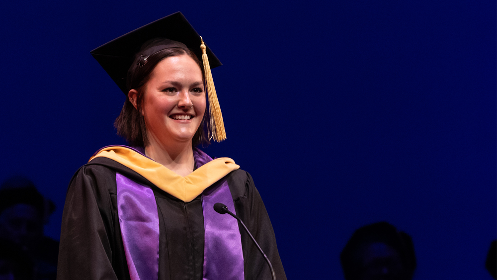 A person wearing a graduation cap and gown smiles on stage in front of a microphone.