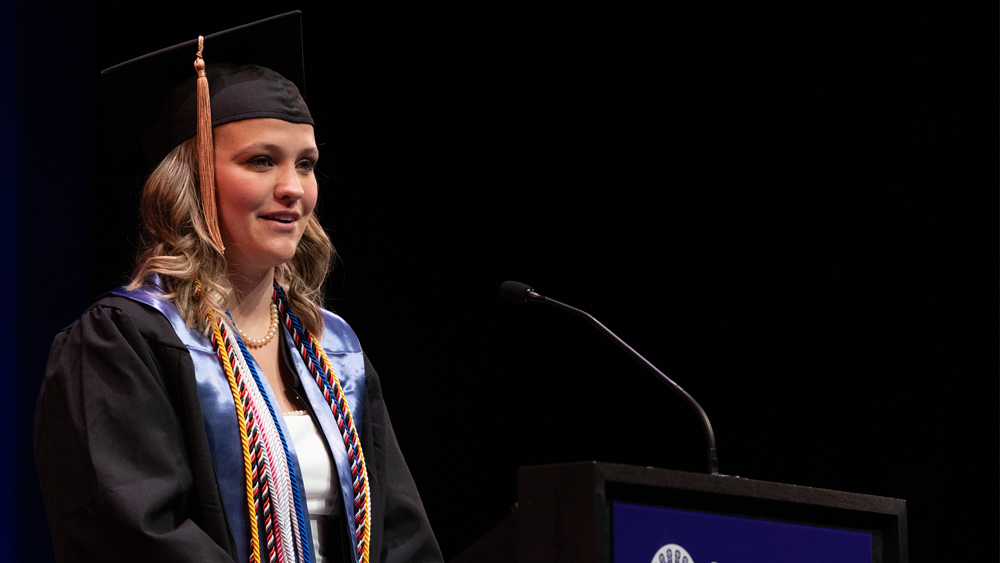 A person wearing a graduation cap and gown with multi-colored cords stands in front of a microphone and podium.