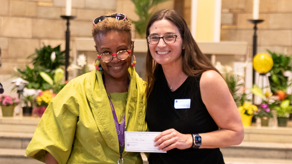 Two women stand in front of an altar and smile; one of them holds a paper check.