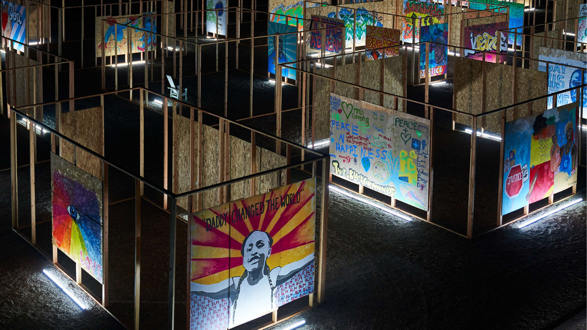A nighttime view of an art installation constructed with wooden frames and partial walls made of plywood. Each wall segment displays colorful, expressive artwork and messages related to social justice, including phrases like "Daddy Changed the World", "I CAN'T BREATHE", and "JUSTICE". Paintings feature portraits, symbolic imagery, and words promoting peace, love, and equality.