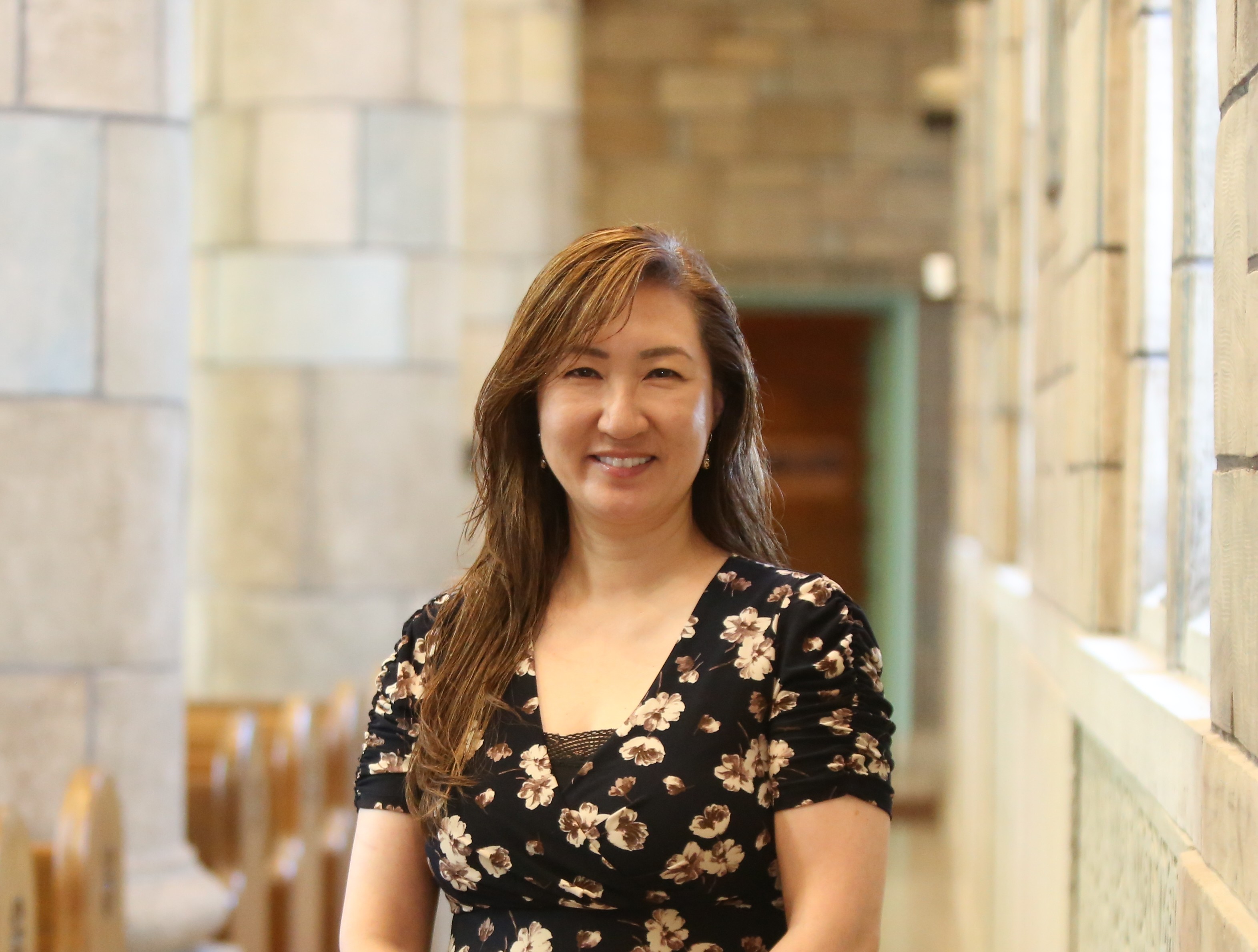 JaeEun Joo, Associate Provost for Adult Education and Online Learning, standing in a stone hallway with wooden pews and arched windows. She is smiling, wearing a black dress with beige floral patterns, and has long brown hair draped over one shoulder.