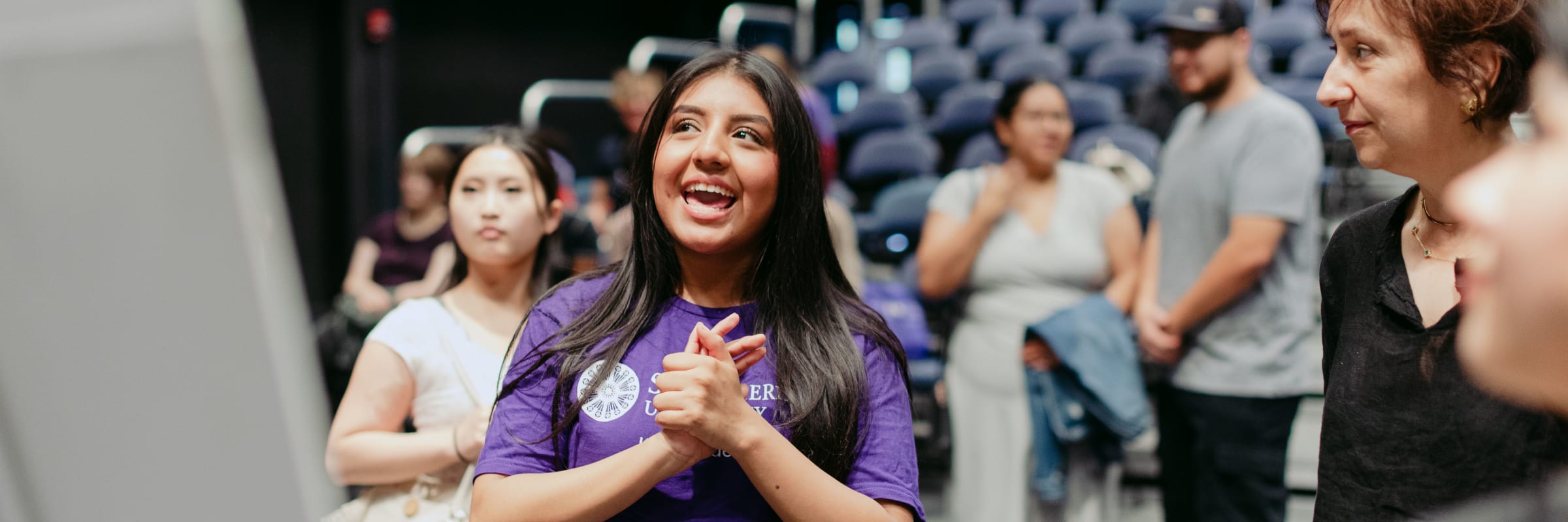 A student in a purple shirt smiles and claps while speaking with others in a seated auditorium setting.