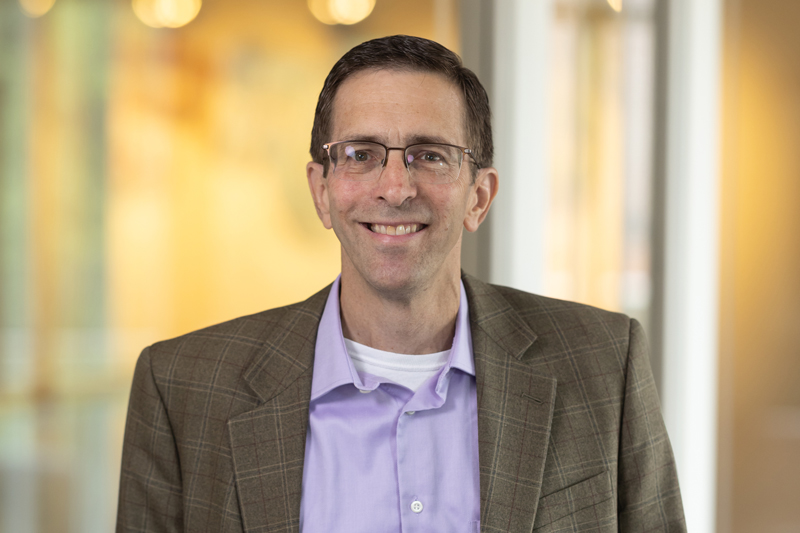 B. J. Miller smiling wearing glasses, a light purple shirt, and a plaid blazer, standing indoors with a blurred background.