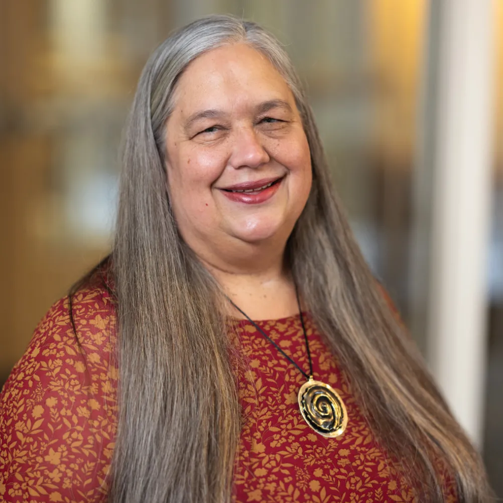 Smiling woman with long gray hair wearing a red and gold patterned top and a large spiral pendant necklace, standing in front of a blurred indoor background.