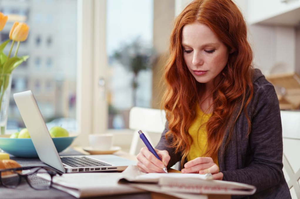 An adult woman online student takes notes while watching a lecture on her laptop.