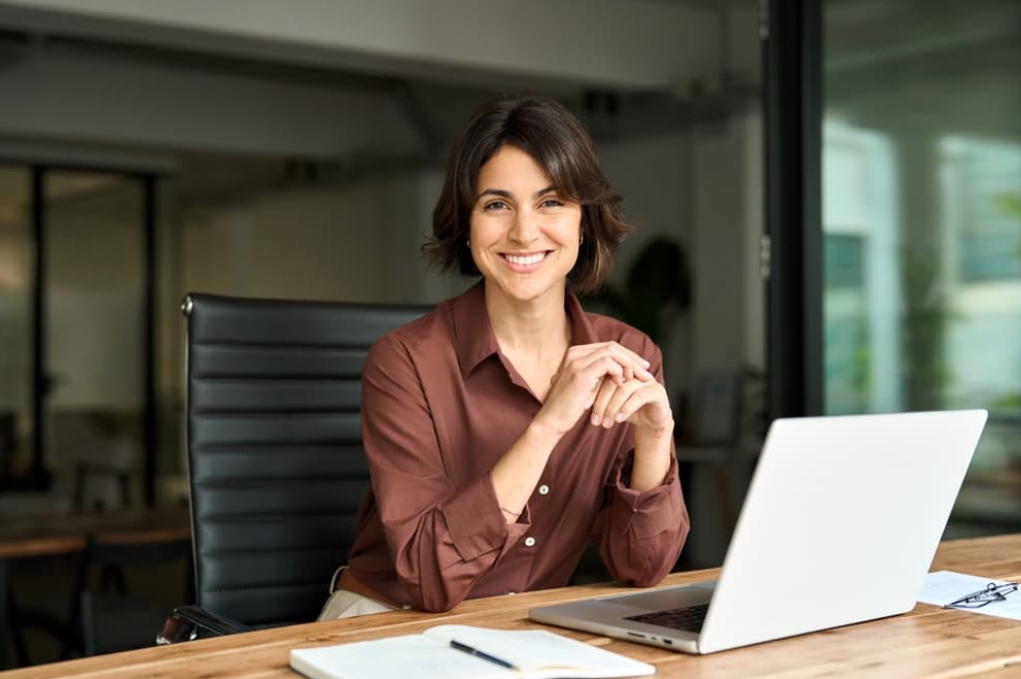  A smiling businesswoman sits working at her desk with a laptop and a paper notebook. 