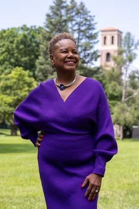Smiling woman in a purple dress standing outdoors on a grassy campus lawn, with trees and a building visible in the background.