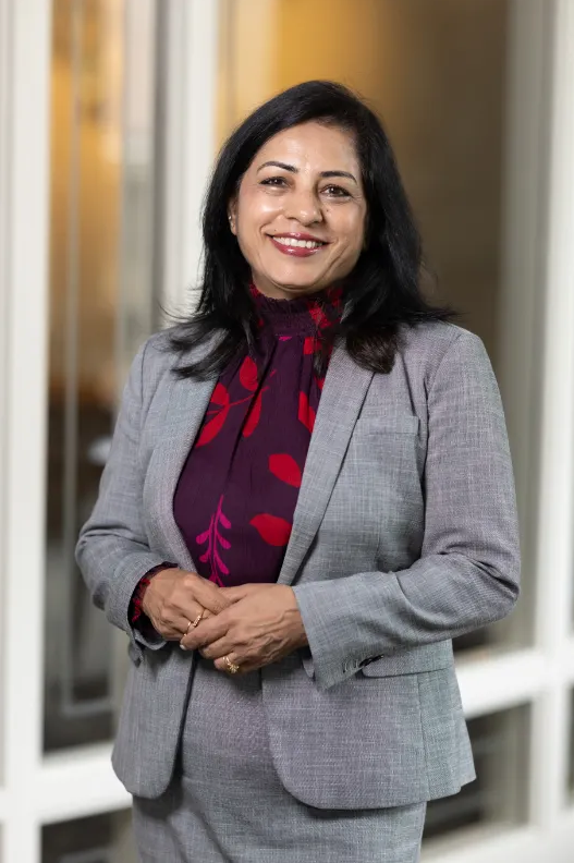 Professional portrait of a smiling woman in business attire, wearing a gray blazer and patterned blouse, standing indoors in front of glass windows with hands clasped.