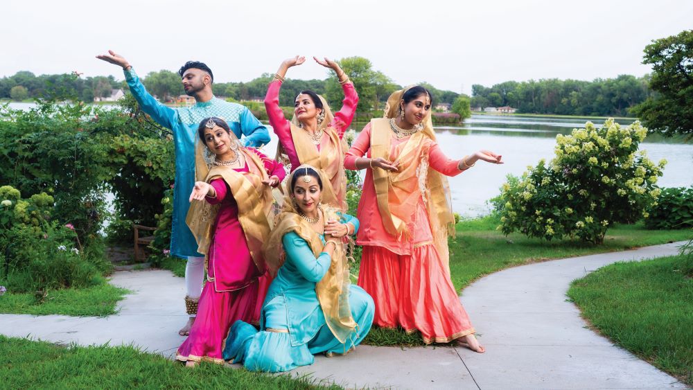 Image of five dancers in colorful costumes against a lush outdoor landscape.