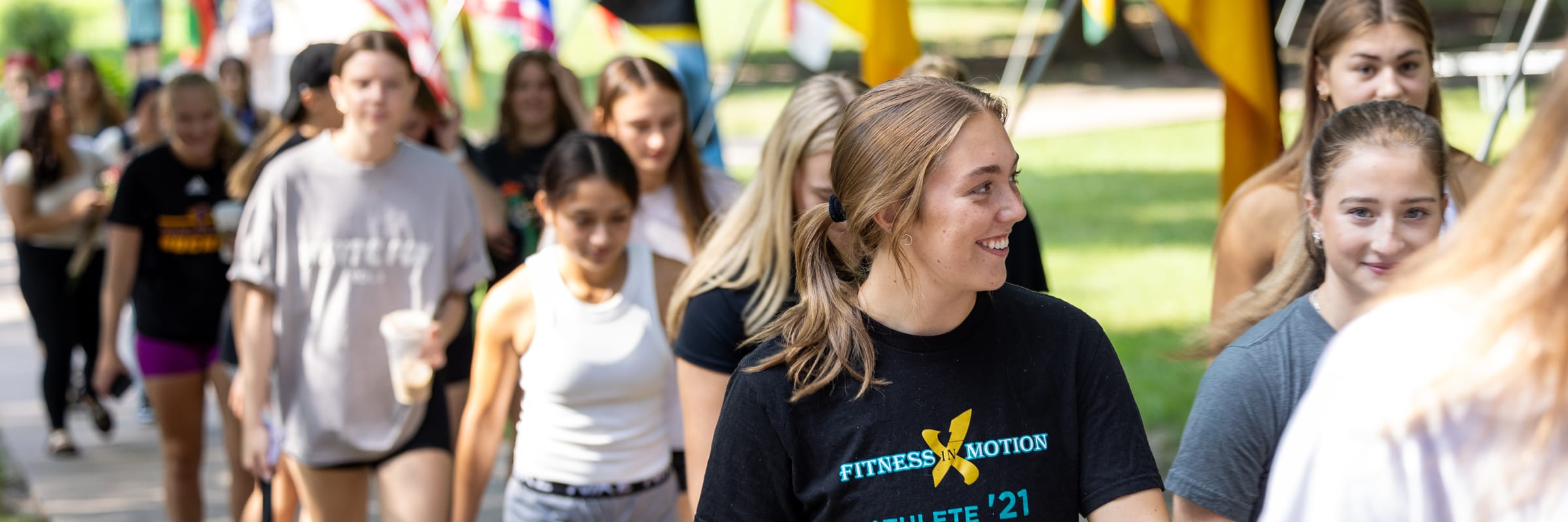 A group of smiling young women walk together outdoors on a sunny day, some carrying drinks, with colorful international flags in the background during a St. Catherine University President’s Forum event.