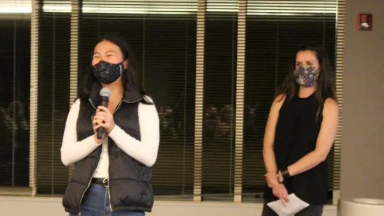 Woman with microphone addresses attendees. Woman stands behind her with paper.