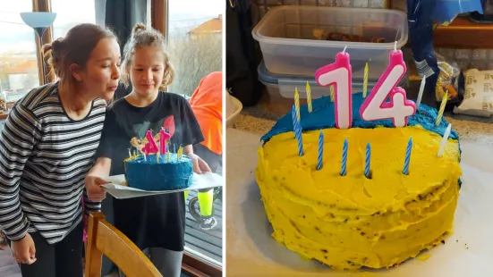 A girl helps her sister blow out a yellow and blue 14th birthday cake