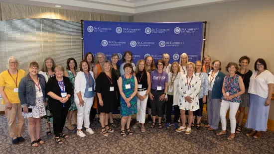 A large group of Reunion celebrants stand together inside, in front of a purple St. Kate's backdrop.