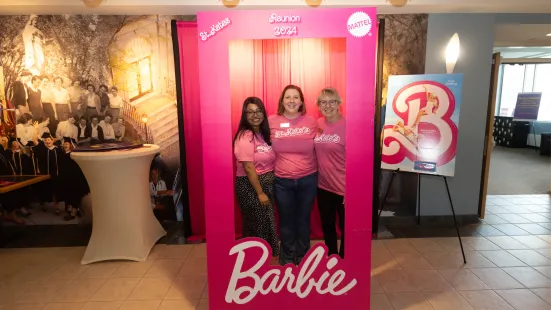 Three women in pink shirts pose in front of a Barbie backdrop.