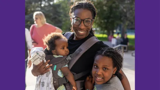 A person wearing classes holds a baby in a carrier to their chest and puts their arm around another child