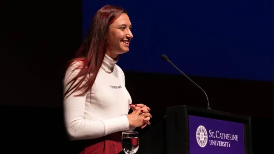 A person with long brown hair wearing a white turtleneck stands in front of a podium.