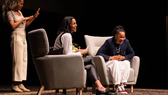 Two women sit on stage in chairs while another one stands behind them interpreting in sign language.
