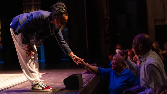 A woman standing by the edge of a stage leans down and reaches out to hold the hand of a person standing beneath the stage. 
