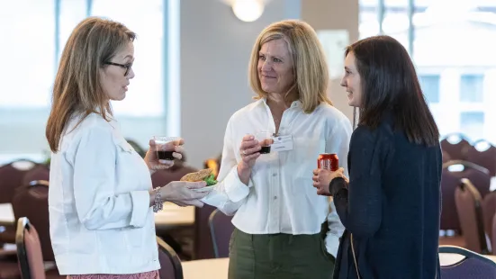 Three smiling women holding drinks stand and talk in a conference room.