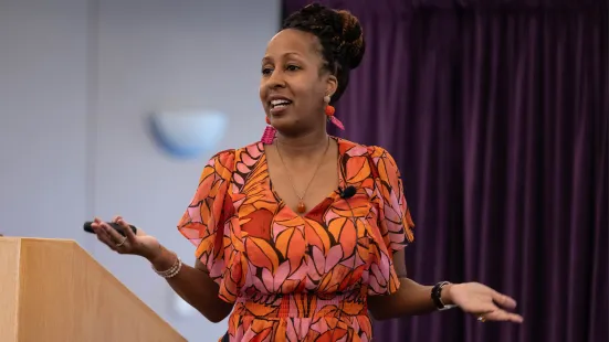 A person wearing a bright orange and pink patterned dress stands in front of a podium, speaking and gesturing their with arms. 
