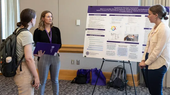Two women stand in front of their poster presentation, talking to a third women wearing a backpack.