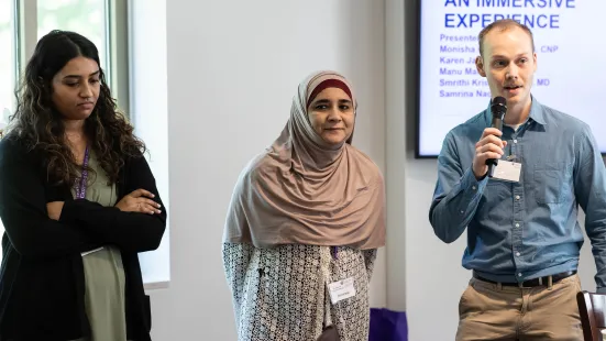 Two women stand next to a man who speaks into a microphone. Behind them in the corner is a powerpoint presentation.