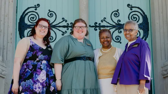Four smiling women stand in front of the turquoise doors to the Chapel.