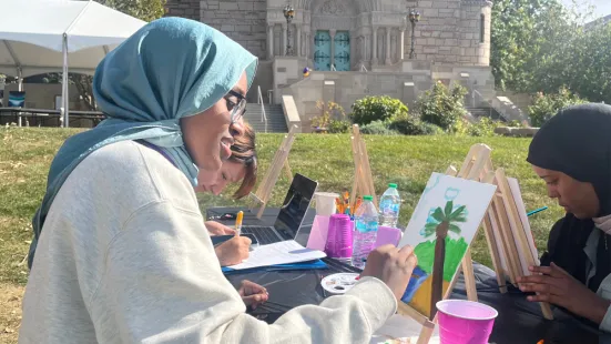 A person sitting outside at a table paints a canvas on a small easel. 