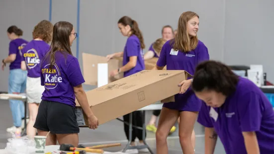 A group of volunteers for Citizen Katie. One student is in the foreground bent over a table. Two students are in the middleground carrying a box. There are four students in the background.