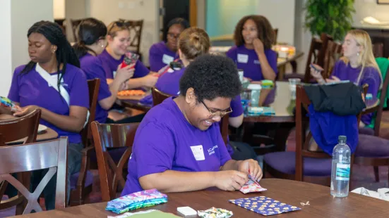 A group of different students volunteering for Citizen Katie. All are sitting at tables wearing purple shirts.