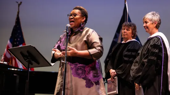 A woman sings into a microphone. On the side, two women in academic robes look on.
