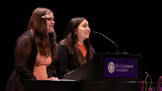 Two students with long hair stand in front of a podium.