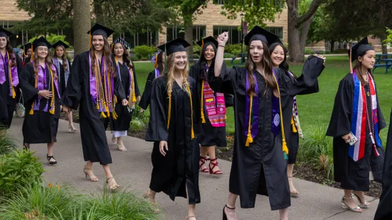 A group of graduates wearing caps and gowns walk outside in a line.