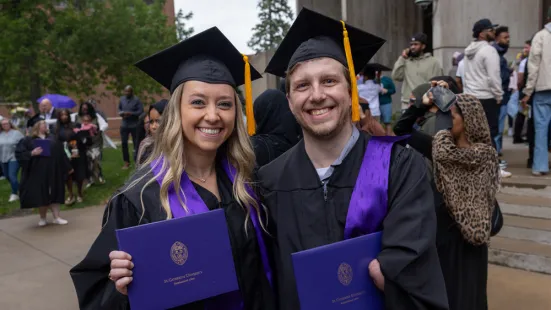 Two people wearing graduation caps and gowns smile and hold up purple diplomas.