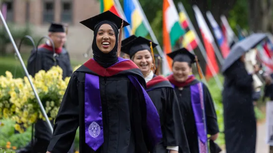A graduate wearing a cap and gown walks down a campus path lined with flags.