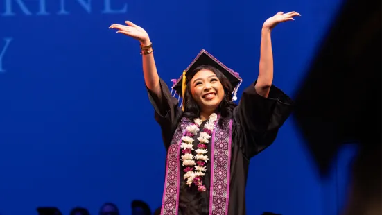A person wearing a graduation cap and gown and standing in front of a blue background lifts their hands up and smiles.
