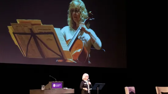 Janet Horvath, MFA, speaks onstage while displaying a photo of herself playing cello onscreen
