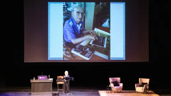 Janet Horvath speaks onstage, displaying a photo of her father onscreen