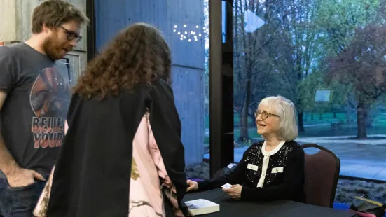 Janet Horvath, MFA, seated behind a table, autographs books while chatting with attendees, standing